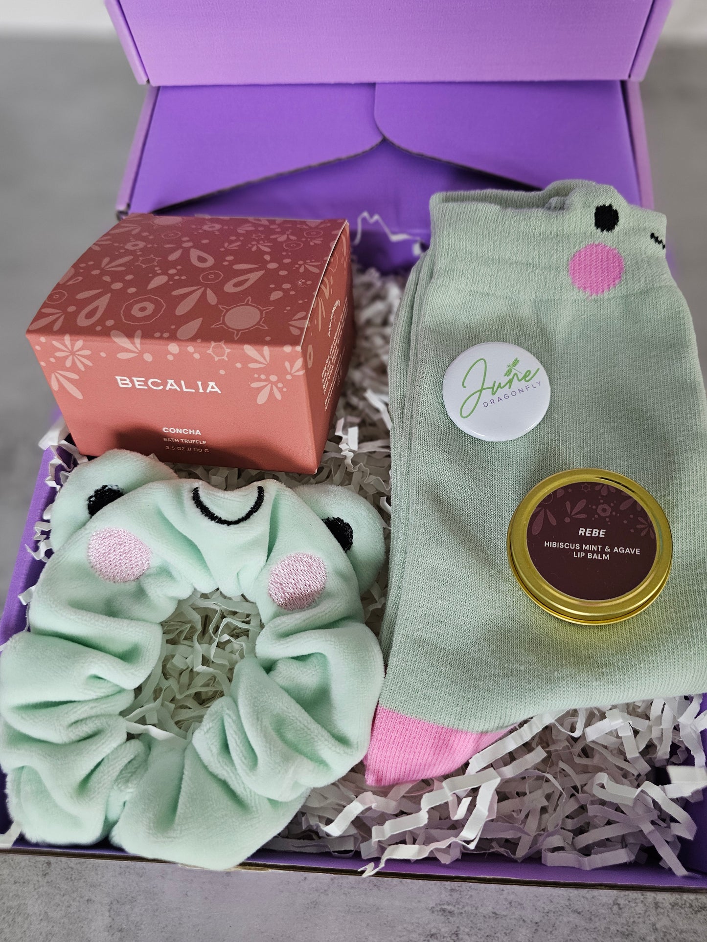 Purple June Dragonfly gift box that features Becalia Botanicals Hibiscus Nourishing Clay Mask, Mint and Apple Froggy Socks, Mint and Apple Froggy Scrunchie, Becalia Botanicals Rebe Hibiscus Mint + Agave Lip Balm and June Dragonfly pin