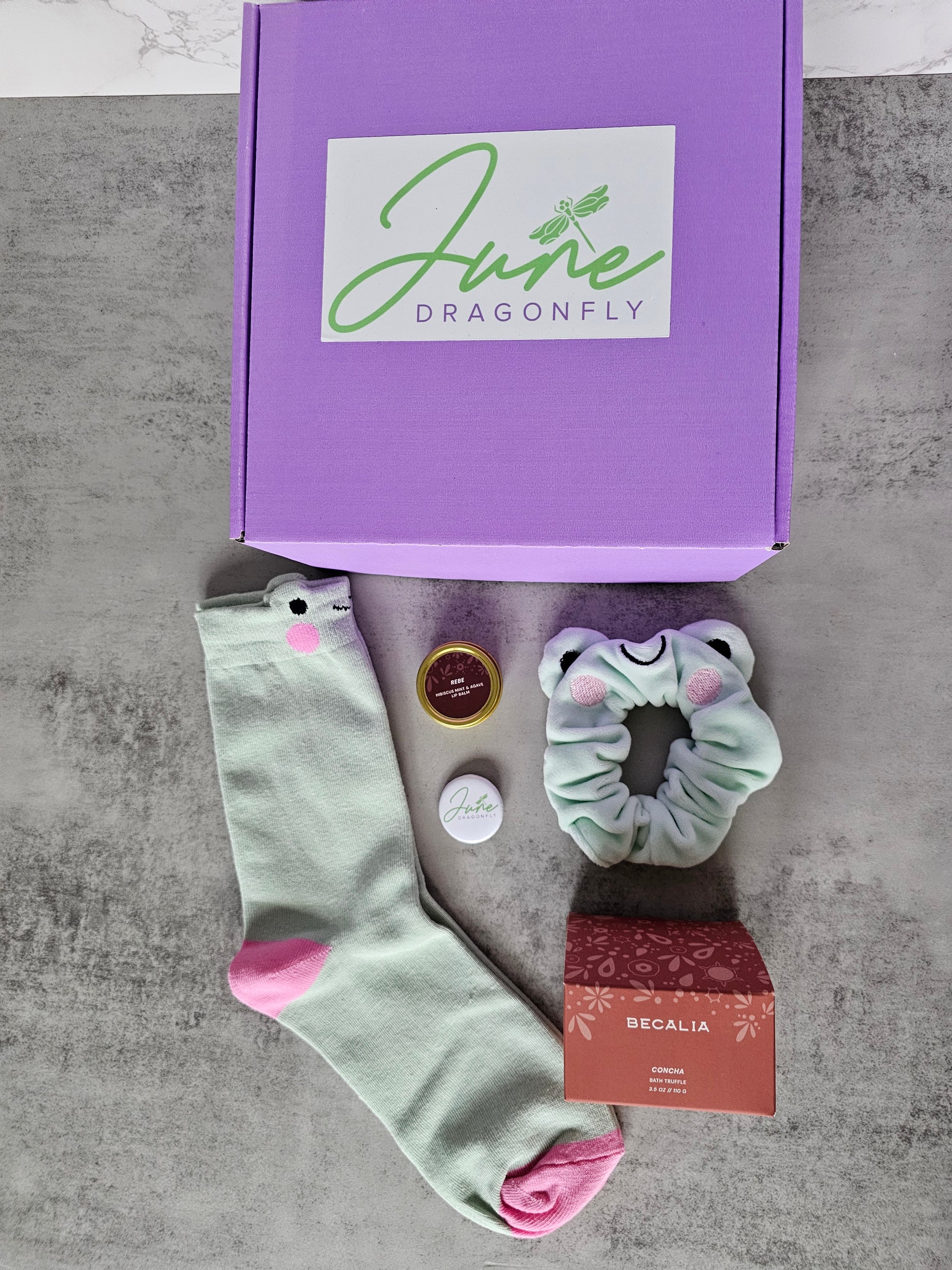 Purple June Dragonfly gift box that features Becalia Botanicals Hibiscus Nourishing Clay Mask, Mint and Apple Froggy Socks, Mint and Apple Froggy Scrunchie, Becalia Botanicals Rebe Hibiscus Mint + Agave Lip Balm and June Dragonfly pin